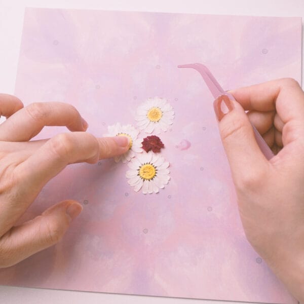 Hands gluing pressed flowers with tweezers onto mandala design sheet, beginner diy step