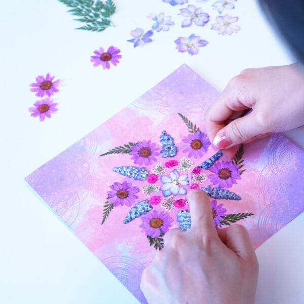 Hands placing pressed flowers on a pink mandala art template as part of a calming DIY ritual