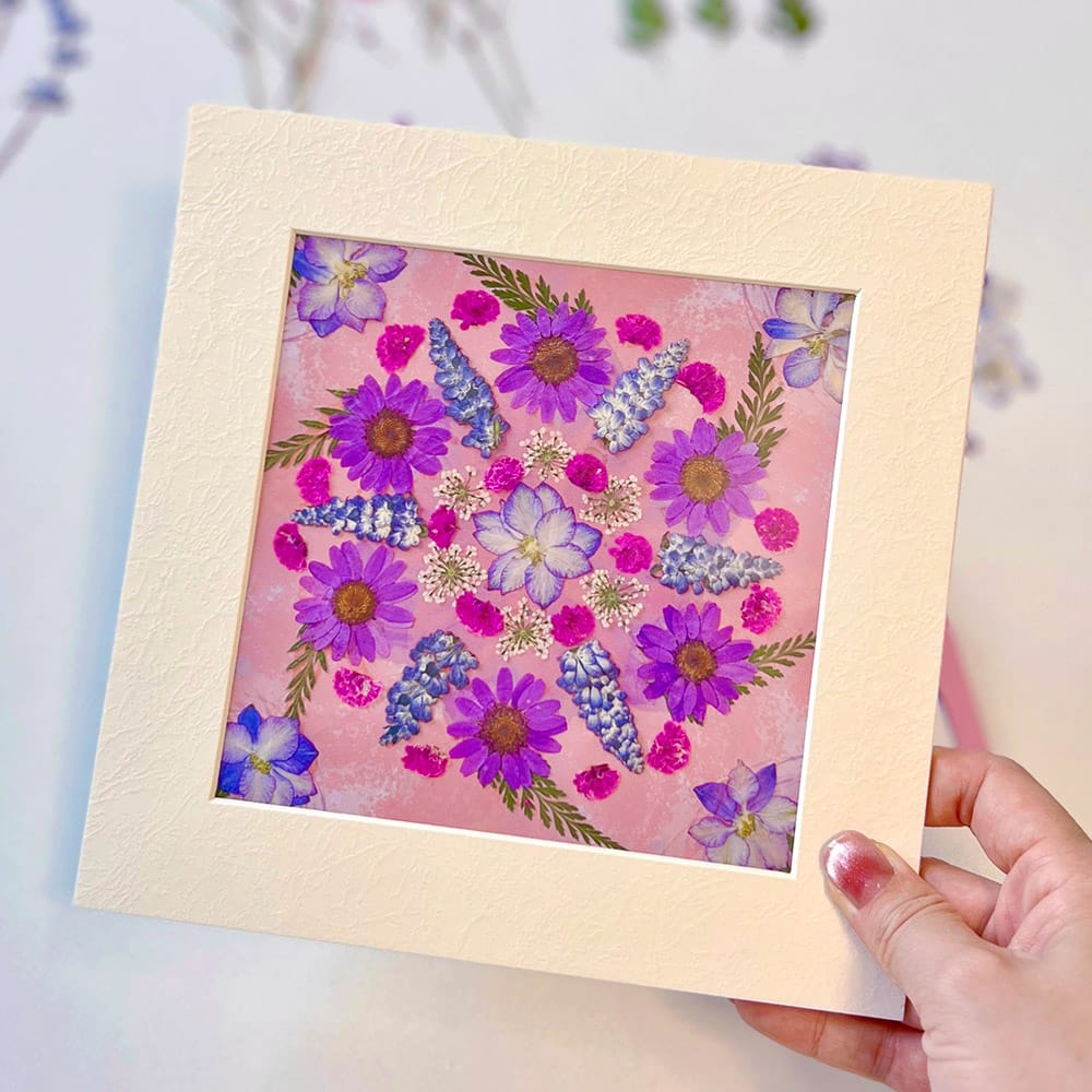 About Us 2 Hand holding a framed pressed flower mandala artwork in soft pink and purple, created with petal and still diy kit.