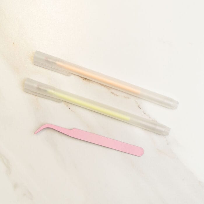 Pressed flower kit tools featuring a pink angled tweezers and two pastel gel pens, used for delicate arrangement of pressed flowers.