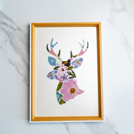 Floral silhouette of a deer head crafted from pressed flowers in soft blues, purples, and pinks on a creamy white background.