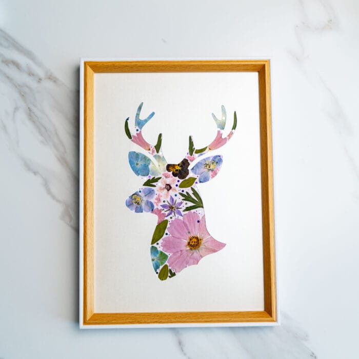 Floral silhouette of a deer head crafted from pressed flowers in soft blues, purples, and pinks on a creamy white background.