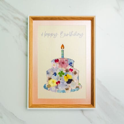 Pressed flower art of a pastel birthday cake silhouette decorated with colorful pressed flowers and a lit candle