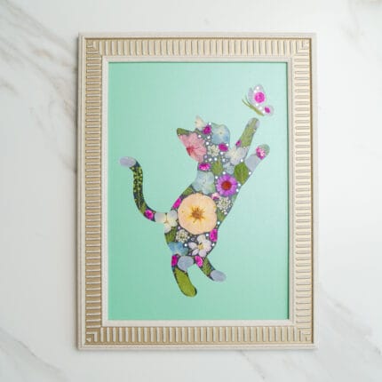 Pressed flower art of a cat silhouette made with flowers, reaching for a butterfly on a green background