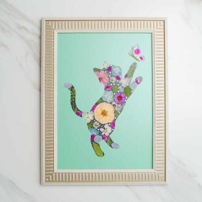 Pressed flower art of a cat silhouette made with flowers, reaching for a butterfly on a green background