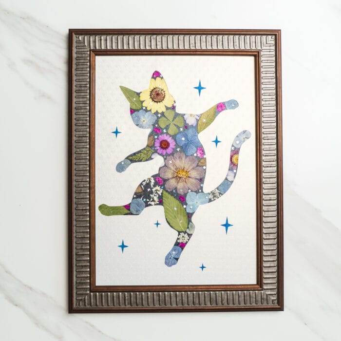 Pressed flower art of a dancing cat silhouette decorated with colorful pressed flowers surrounded by blue stars