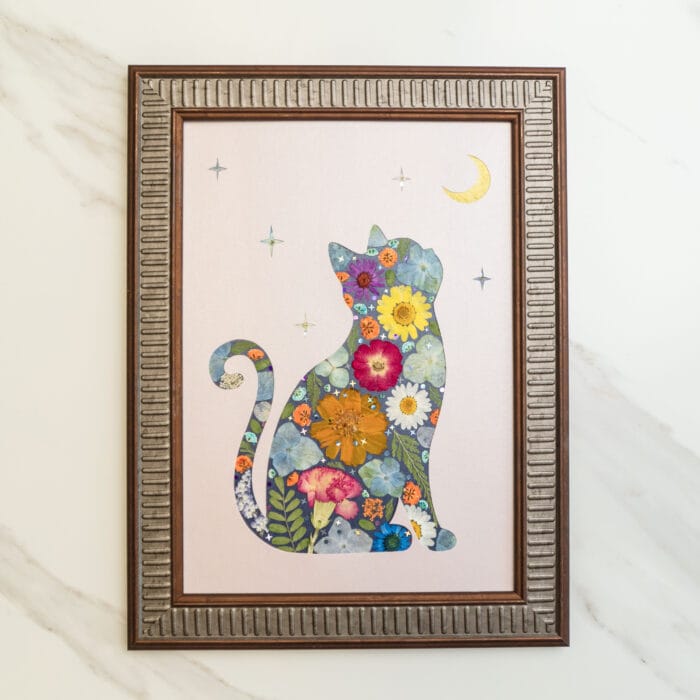 Pressed flower art of a cat silhouette filled with colorful flowers, looking at the moon and stars on a light pink background