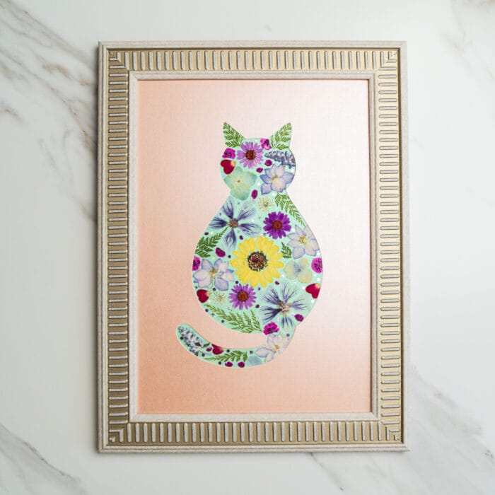 Pressed flower art of a cat silhouette decorated with yellow, purple, and blue flowers on a peach background