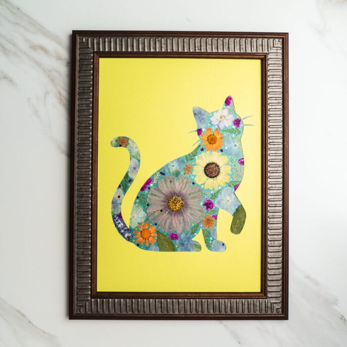 Pressed flower art of a cat silhouette with large pressed flowers in blue and orange on a bright yellow background