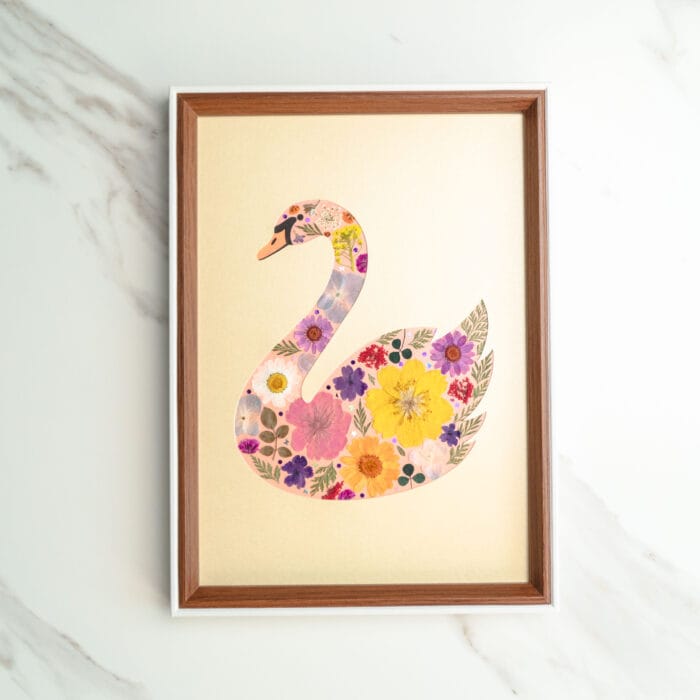 Pressed flower art of a swan silhouette filled with pink, yellow, and purple flowers on a golden background