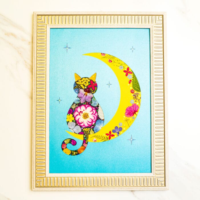 Pressed flower art of a colorful cat silhouette sitting beside a yellow crescent moon with floral details, on a blue background in a decorative frame.