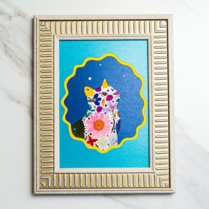 Pressed flower art of a cat silhouette framed in silver, with colorful petals on a blue night sky background.