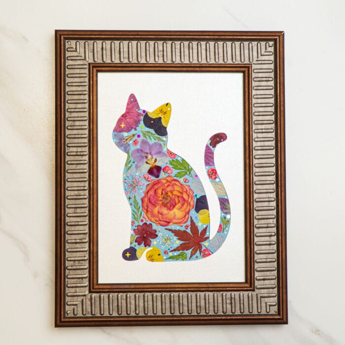 Pressed-flower art of a colorful cat silhouette, framed with a textured pinkish-brown border, featuring vibrant petals and leaves on a light background.