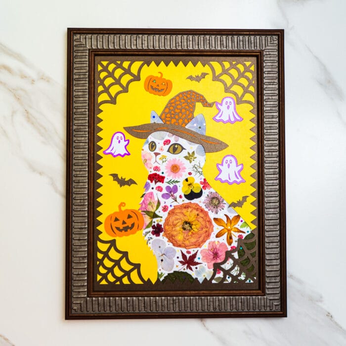 Pressed flower halloween cat artwork with witch hat, pumpkins, bats, and ghost decorations in a brown frame on yellow background.