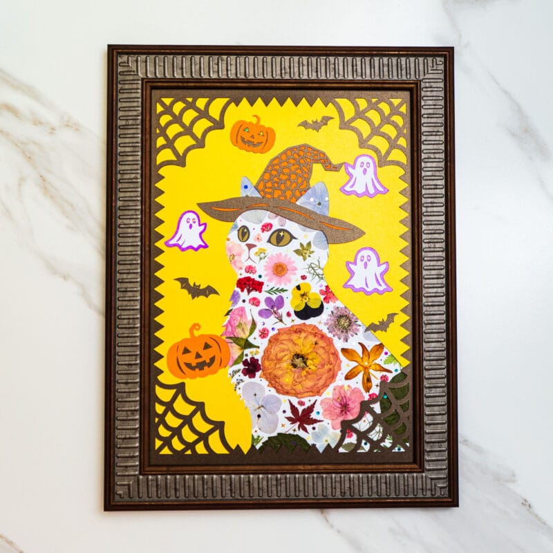 Pressed flower halloween cat artwork with witch hat, pumpkins, bats, and ghost decorations in a brown frame on yellow background.