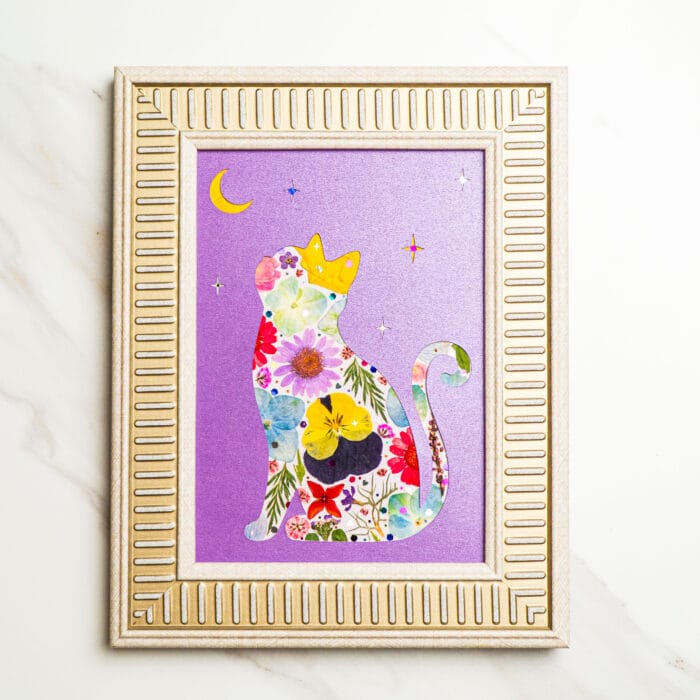 Pressed flower art cat silhouette decorated with colorful blossoms, wearing a golden crown and looking at the moon, on a purple background with stars, framed in light gold frame.