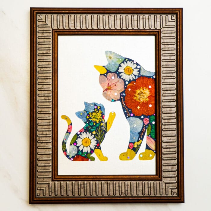 Pressed flower art showing a mother cat and kitten silhouette filled with colorful petals, in a bronze frame.