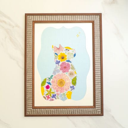 Pressed flower art cat silhouette with pastel blooms on a soft blue sky background, gazing at stars, framed in vintage brown frame.