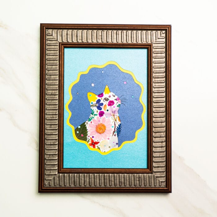 Pressed flower cat silhouette decorated with vibrant blossoms, set against a blue background with yellow scalloped border.