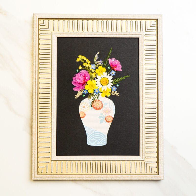 Pressed flower bouquet in a patterned vase on black background, framed in silver.